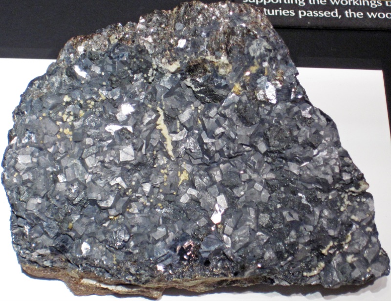 A slab of Laurion ore on museum display: dark grey galena made of many small, glinting cubic crystals, flecked with pale mineral grains, on a brownish rock base.