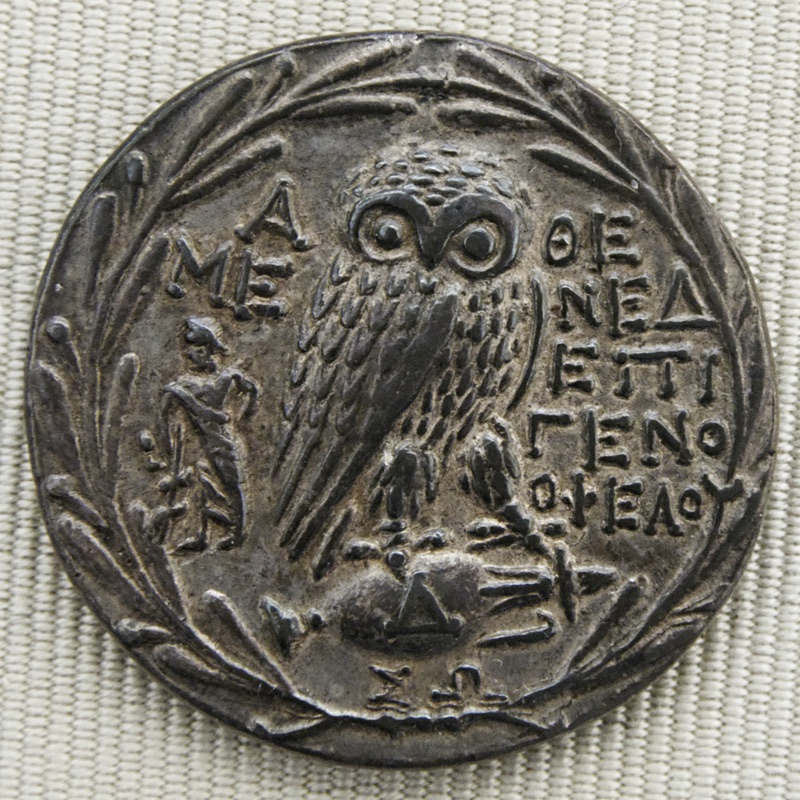 A New Style owl stands on a horizontal amphora amid inscriptions, inside an olive wreath.